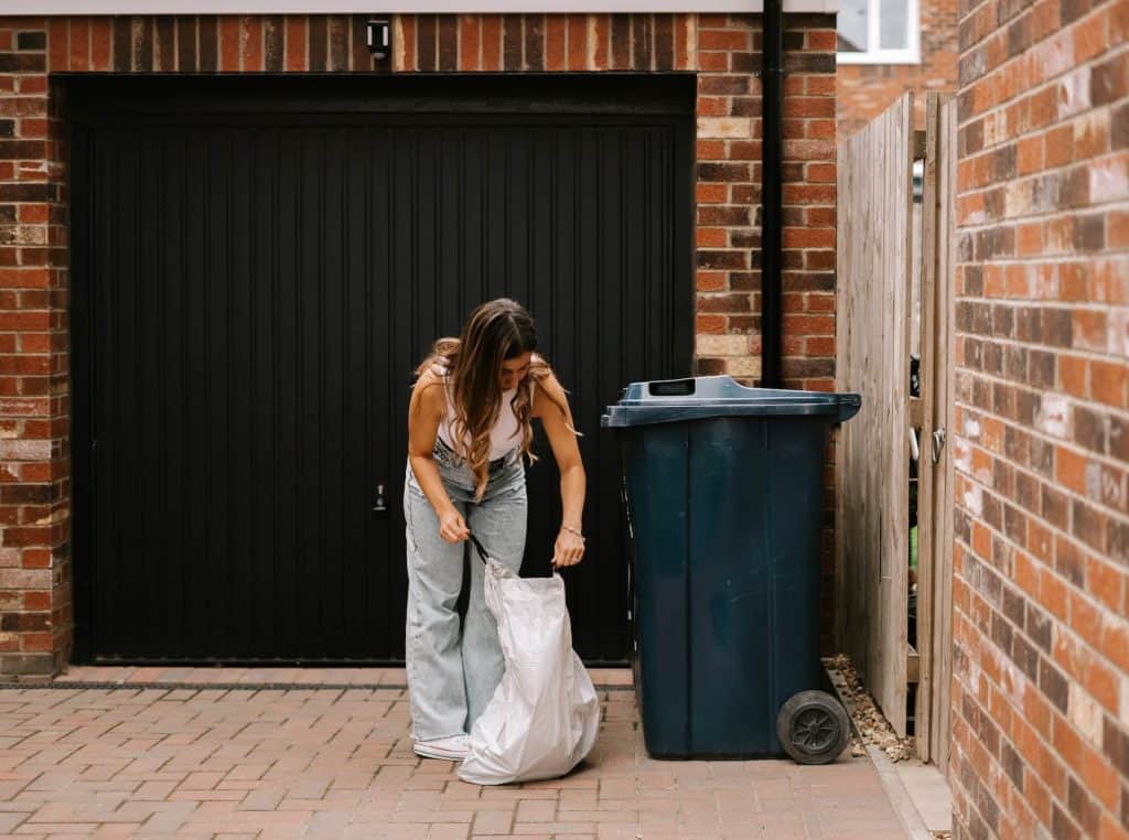 A person wearing a striped top and jeans is crouching in front of a black garage door, tying a white garbage bag next to a blue trash bin on the brick-paved driveway. Nearby, an Electronics Drop Off Box Atlanta GA stands ready for responsible disposal.