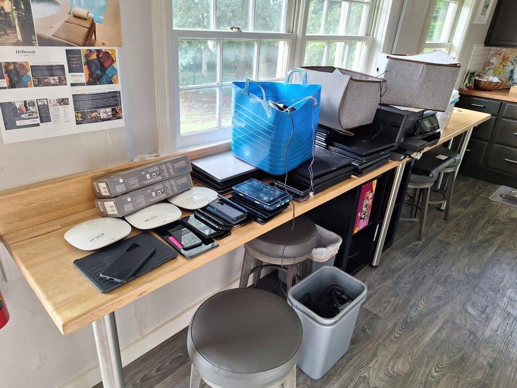 A wooden table against a window holds various electronic devices, including laptops, tablets, and smartphones, awaiting e-waste disposal. Stacks of CDs/DVDs and cables accompany them. Two bins and stools are beneath the table, while a wall is decorated with photos and papers.