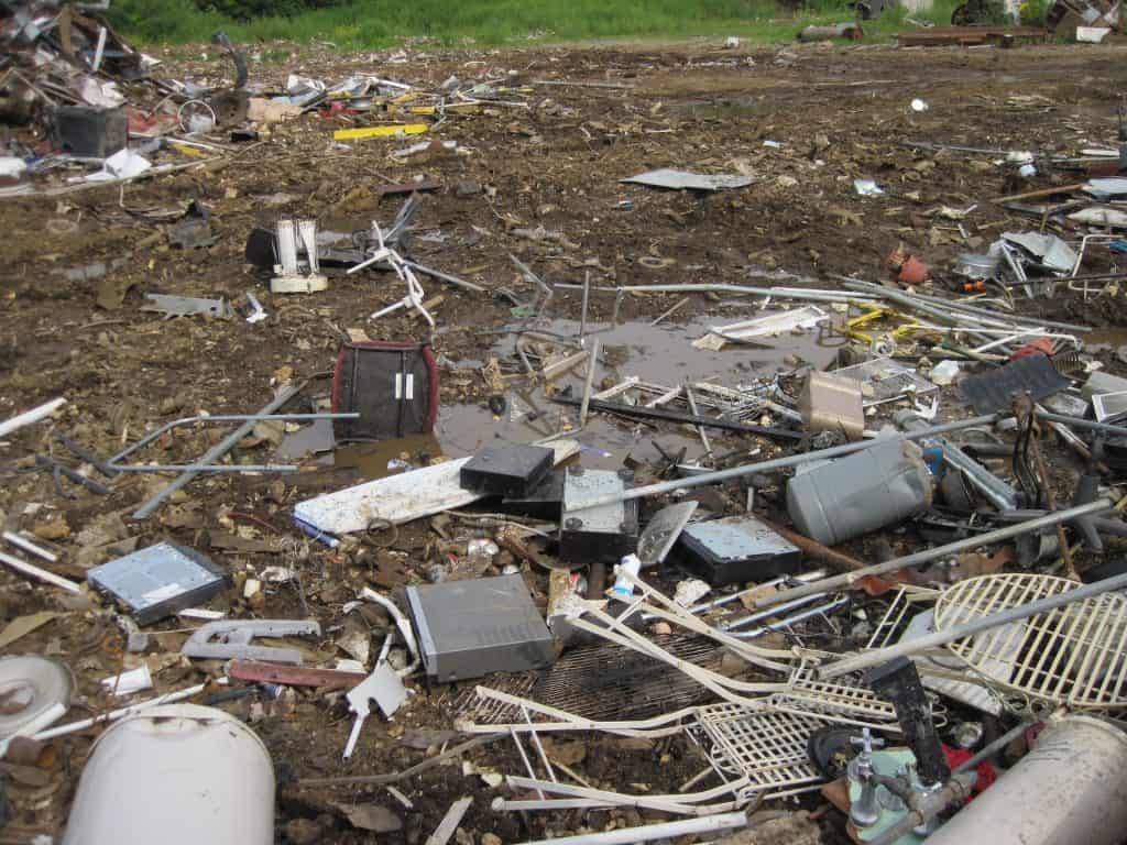 A cluttered junkyard with scattered electronic waste and metal debris sits in stark contrast to the lush green grass beyond. Here in Atlanta, GA, various components like wires and small appliances await their fate, emphasizing the city's need for effective electronic recycling solutions.