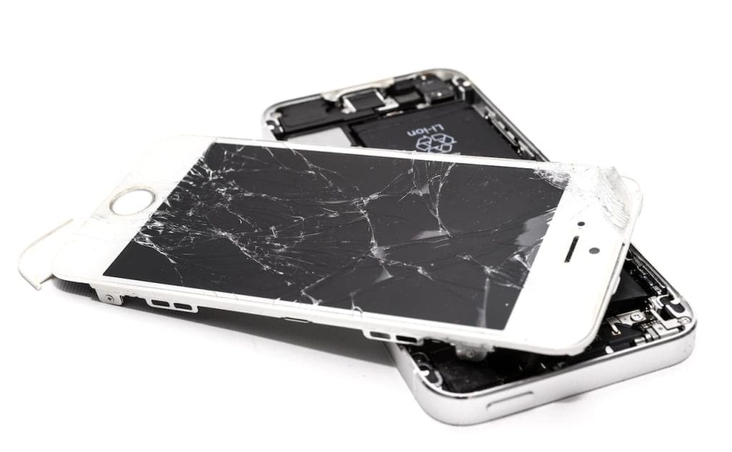 Two old smartphones with shattered screens are stacked on top of each other against a white background. The top phone's screen is extensively cracked, displaying the phone's inner components partially exposed below. Consider using an electronic recycling facility in Atlanta, GA, to dispose of them responsibly.