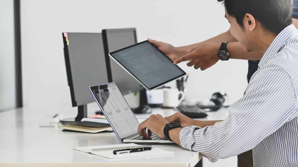 Two people are working in an office. One is typing on a laptop, and the other is holding a tablet with code displayed. Both are seated at a desk with computer monitors, a notebook, and a pen nearby, discussing plans for setting up an electronic recycling drop-off box in Atlanta, GA.