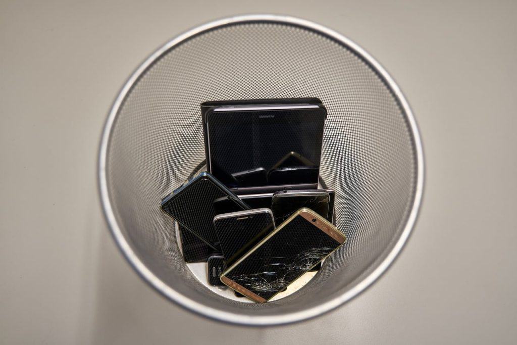 A top-down view of a metal trash can resembling a Drop Off Box in Atlanta, GA, filled with multiple discarded smartphones, some with cracked screens, on a light-colored floor.
