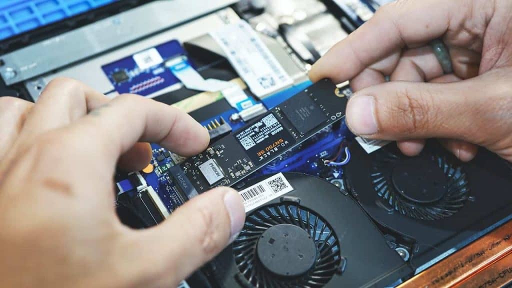 A close-up of hands installing an SSD into a laptop's internal compartment highlights the precision needed in aligning the electronics. Visible components include cooling fans, memory slots, and circuit boards, reminiscent of expert repairs at Electronics Atlanta GA.