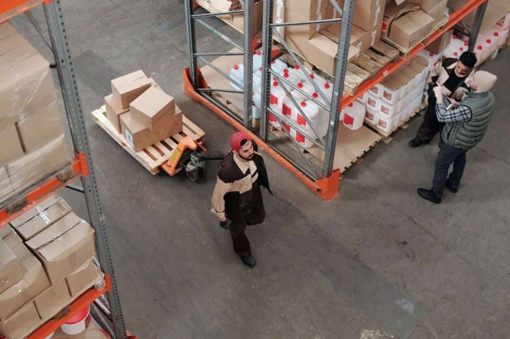 A man in a brown jacket and red cap walks through a warehouse in Atlanta, GA, pulling a cart loaded with cardboard boxes. Two other men discuss electronic recycling in the background, surrounded by shelves stocked with various goods.