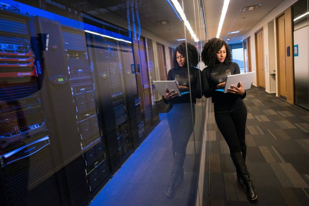 A woman stands in a hallway next to a glass wall reflecting server racks, focused on her laptop. The setting is a sleek, modern office, emphasizing tech-forward solutions like electronic recycling.