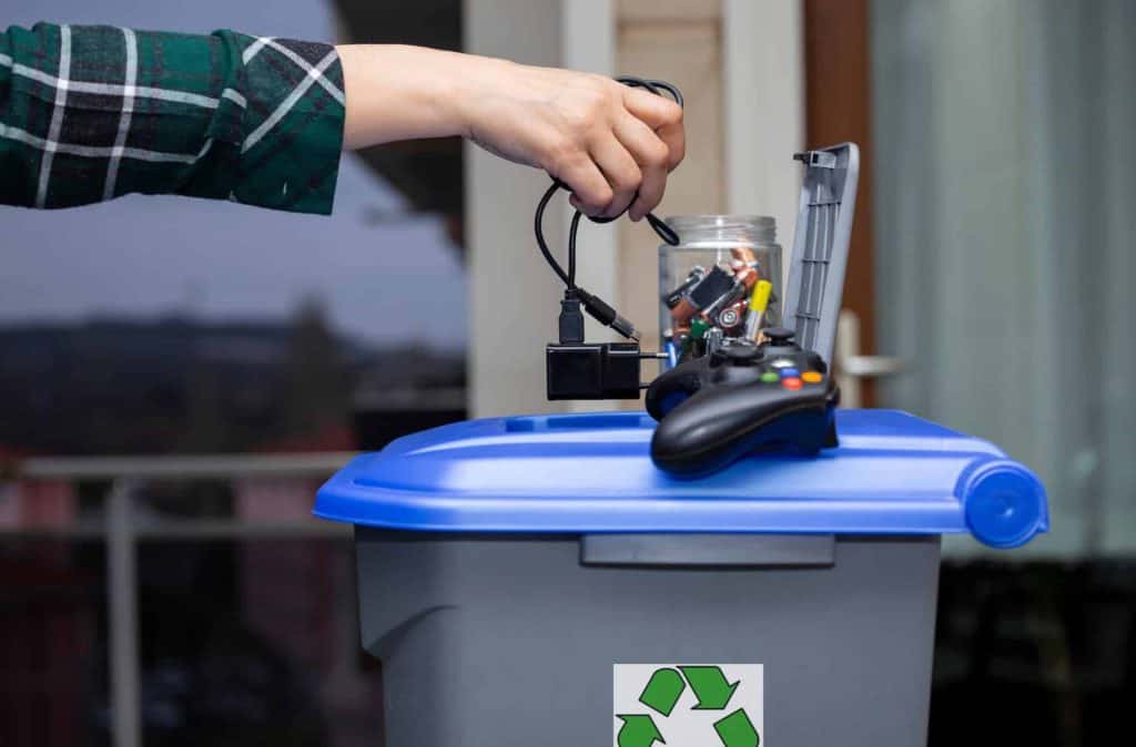 A person in Atlanta, GA, places electronic waste—cables, batteries, and a game controller—into a blue-lidded recycling bin marked with the iconic recycling symbol. The background reveals a blurred outdoor scene that suggests an Electronic Recycling drop-off box location.