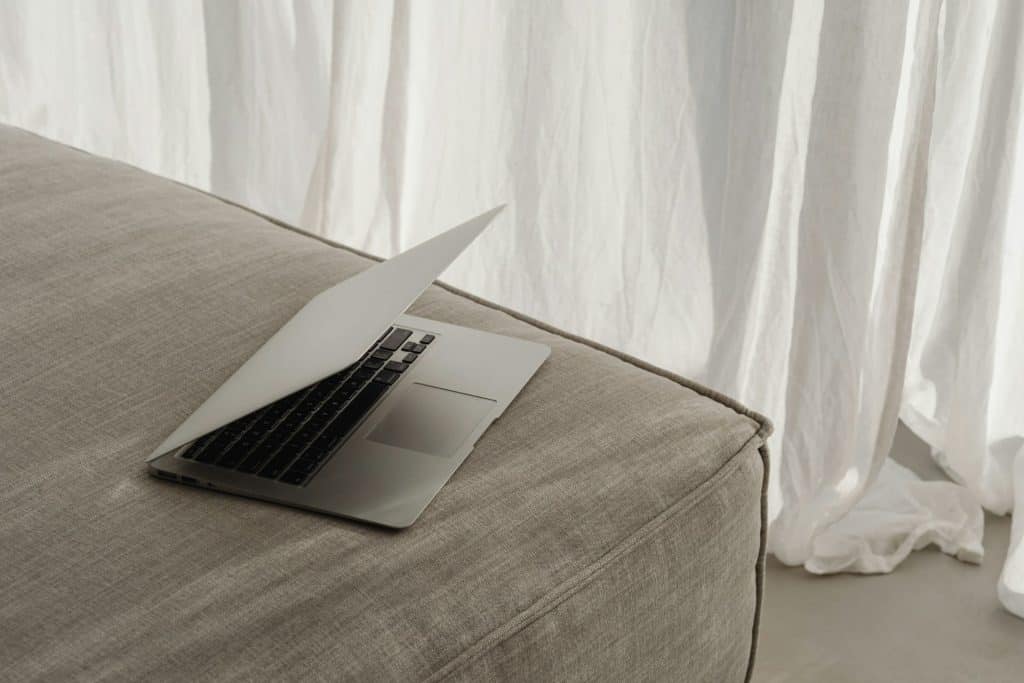 A partially open laptop rests on a beige fabric sofa against a backdrop of white, flowing curtains. Subtly lit, the scene exudes calm and minimalism, echoing the importance of electronic recycling in Atlanta, GA for a sustainable future.