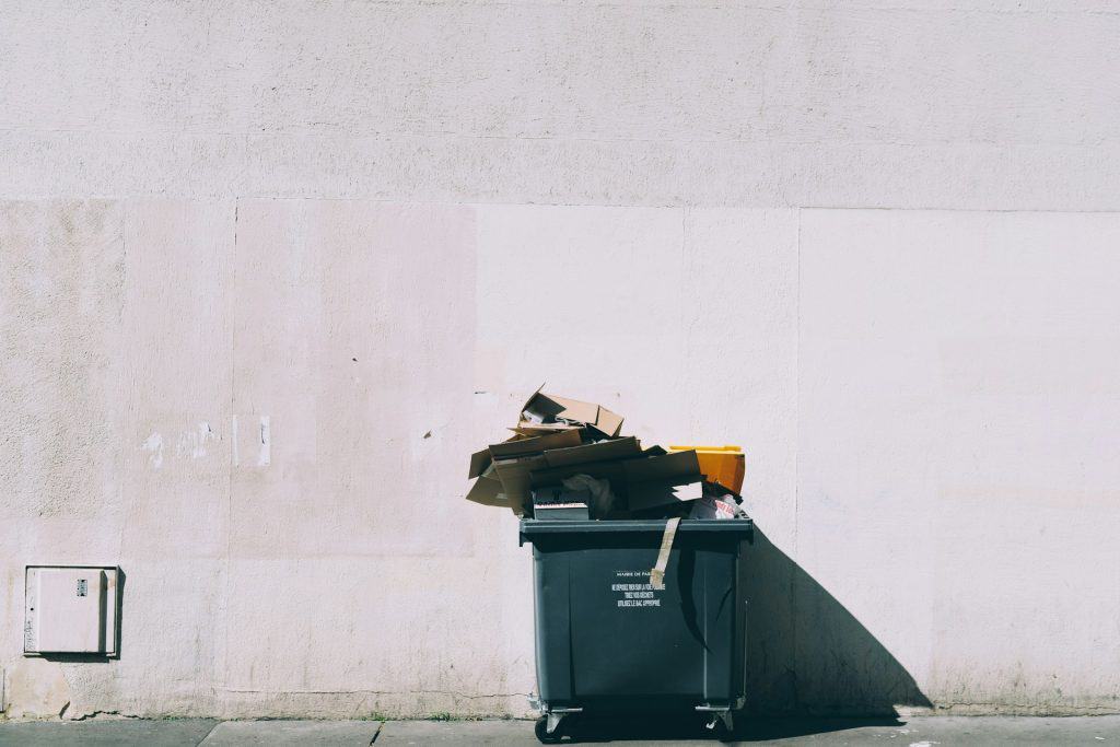 A large, overflowing dumpster with cardboard boxes and trash on top stands against a blank, white wall. Nearby, an electronic recycling drop-off box invites residents of Atlanta, GA to discard responsibly. The shadows on the wall indicate a sunny day, and the ground is paved with concrete.
