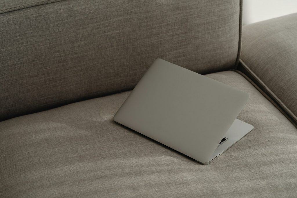 A partially closed silver laptop sits on a gray fabric couch, angled slightly with the keyboard barely visible. The background shows the couch's cushion and armrest, embodying a minimal and cozy atmosphere. Consider electronic recycling in Atlanta, GA to ensure your unused devices find new life.