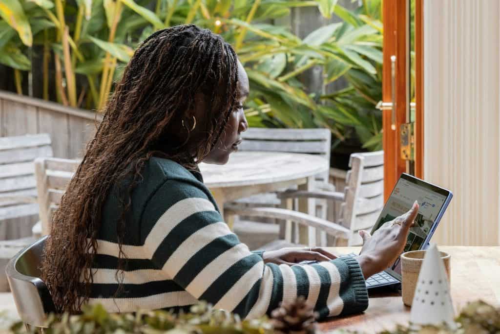 A person with long hair and a striped sweater is sitting at a table, using a laptop in the serene garden setting. Nearby, a drop-off box for electronic waste in Atlanta, GA encourages eco-friendly habits amidst the lush greenery and wooden furniture.