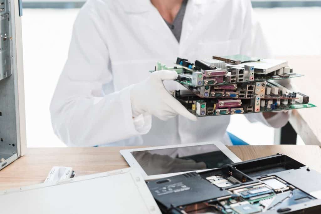 A person in a white lab coat and gloves holds several computer circuit boards, seated at a desk with electronic components. Nearby, an open laptop displays its internals. This scene highlights the crucial role of electronic recycling in Atlanta, GA.