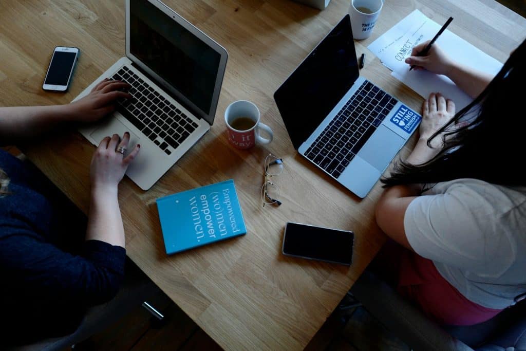 Two people working on laptops at a wooden table in Atlanta, GA, with notebooks, smartphones, and coffee mugs. A pair of glasses and a blue book titled "empowered women empower women" are also on the table. Nearby, a drop-off box for electronic waste awaits contributions.