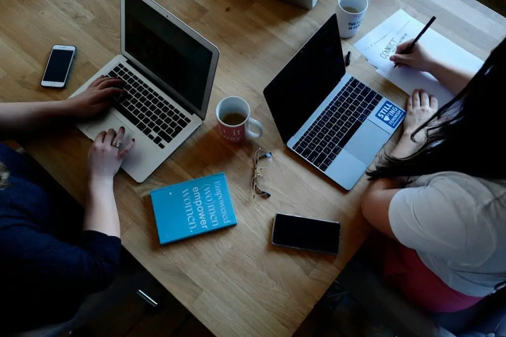 Two people working on laptops at a wooden table in Atlanta, GA, with notebooks, smartphones, and coffee mugs. A pair of glasses and a blue book titled "empowered women empower women" are also on the table. Nearby, a drop-off box for electronic waste awaits contributions.