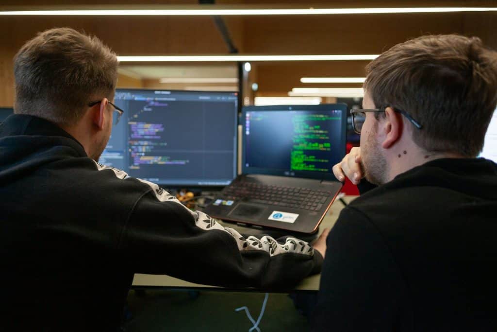 Two people are seated at a desk, facing computer monitors displaying code. One uses a laptop, and the other looks at a larger screen. Both wear glasses and are focused on programming in their modern office in Electronics Atlanta GA.
