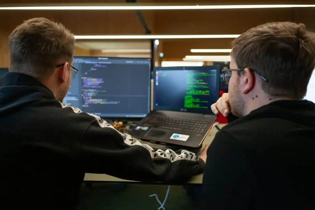 Two people are seated at a desk, facing computer monitors displaying code. One uses a laptop, and the other looks at a larger screen. Both wear glasses and are focused on programming in their modern office in Electronics Atlanta GA.