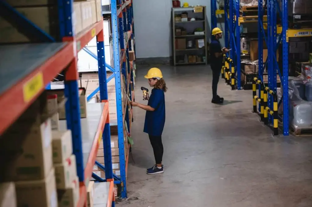 A person wearing a yellow hard hat and blue shirt uses a scanner to check inventory on a warehouse shelf filled with electronics. Another person, also in work attire, is seen in the background. The warehouse, part of an electronic recycling facility in Atlanta, GA, is organized with blue shelving units.