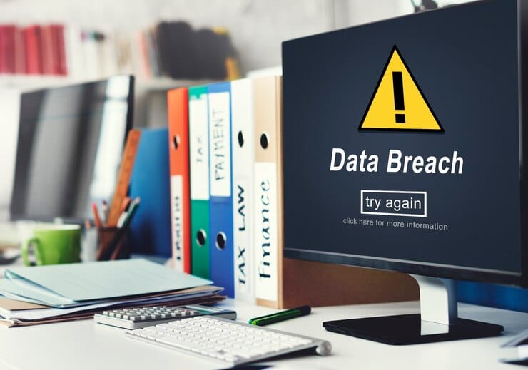 A computer screen in an Atlanta office flashes a "Data Breach" warning with a yellow caution symbol. Below, a "try again" button awaits action. Nearby, folders labeled "TAX," "LAW," and "FINANCE" are surrounded by office supplies amid thoughts of electronic waste disposal options.