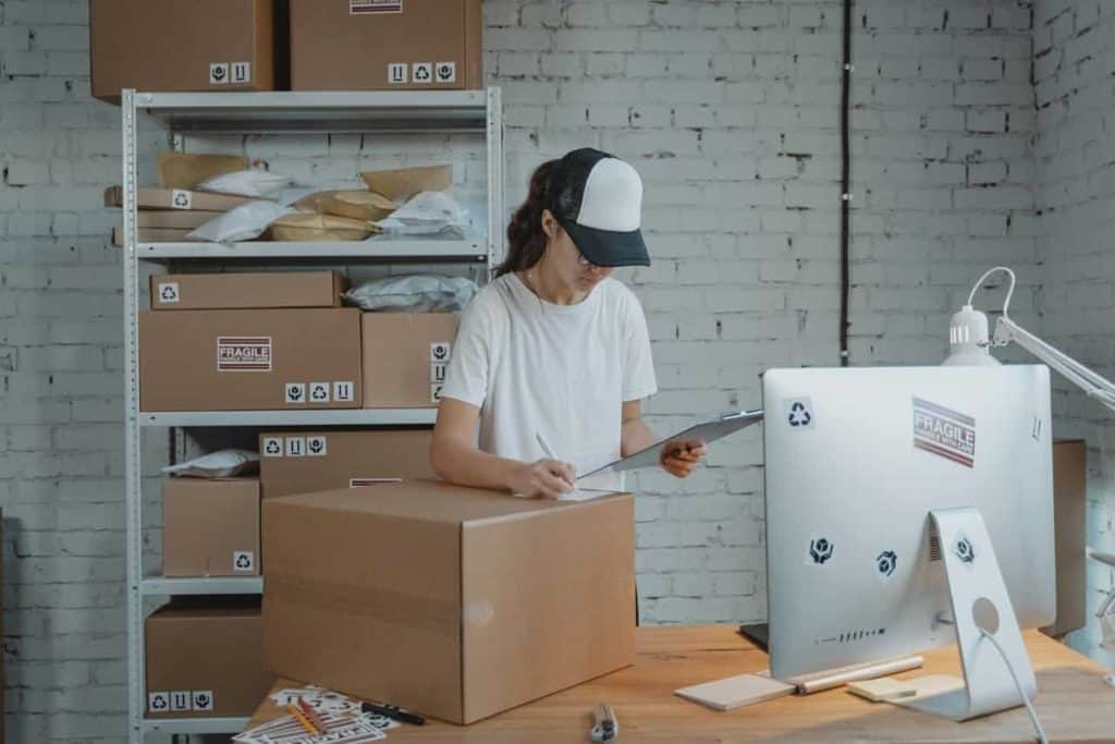 A person in a white shirt and cap stands at a desk in a warehouse, checking a clipboard. They are surrounded by large and small cardboard boxes for electronics recycling in Atlanta, GA, with a computer and lamp on the desk. The white brick wall adds to the industrial feel.