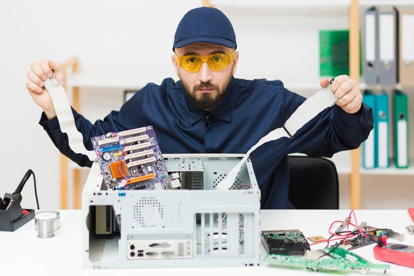 A person with a beard, wearing a cap and yellow protective glasses, sits at a desk in Atlanta, GA, assembling a computer tower. They hold two ribbon cables, surrounded by scattered computer parts and tools, emphasizing the city's commitment to reducing electronic waste.