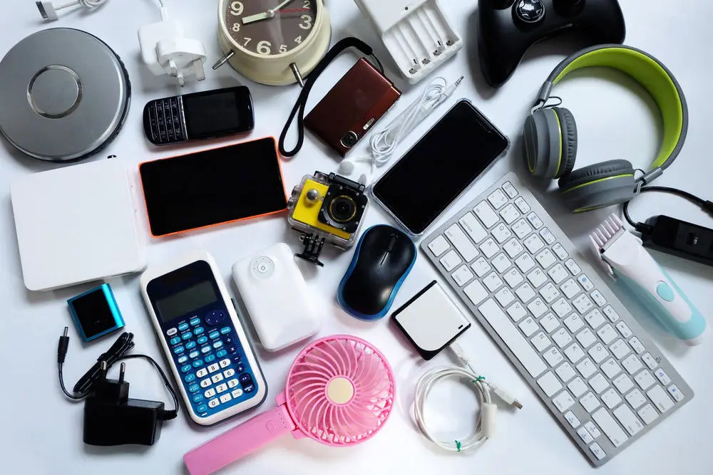 A collection of electronic gadgets, including a camera, headphones, smartphone, keyboard, gaming controller, calculator, mouse, and various chargers arranged on a white surface near an Electronic Recycling Drop Off Box in Atlanta GA.