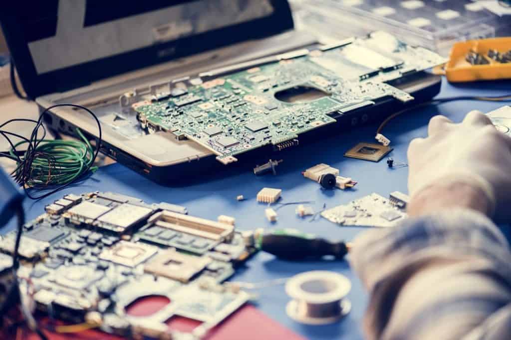 A person works diligently on dismantling and repairing laptop components on a table. Various electronic parts, wires, and tools are scattered around as a gloved hand holds a small component from the partially disassembled laptop. This scene highlights the importance of addressing electronic waste in Atlanta, GA.