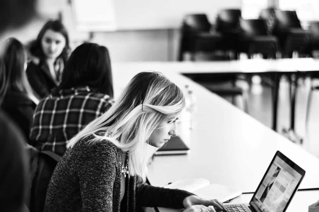A black and white image captures a woman with light hair working on a laptop at a desk. In the background, others casually chat, possibly discussing topics like electronic recycling in Atlanta, GA. The setting conveys a relaxed meeting or study session atmosphere.