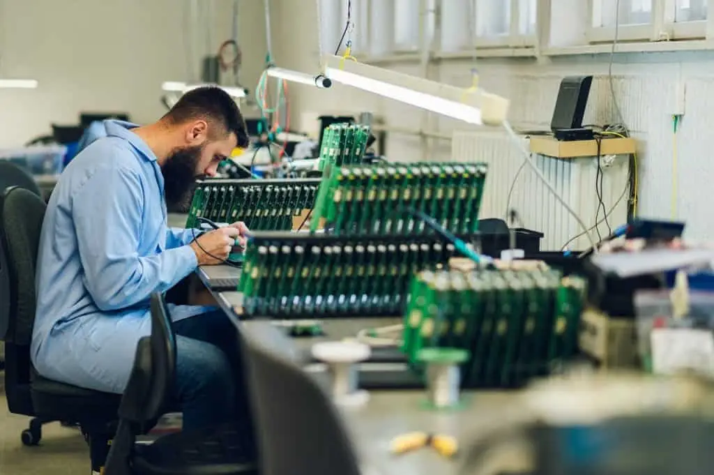 A technician in a blue lab coat is meticulously assembling circuit boards at a well-organized workstation in Atlanta, GA. Surrounded by various electronic components and equipment, the scene highlights the city's pivotal role in electronics innovation.