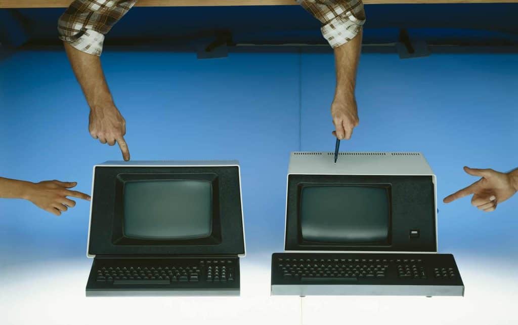 Two vintage computers are positioned side by side, with hands pointing at each one, against a blue background. Each monitor has a keyboard in front. These relics remind us of the growing pile of electronic waste in Atlanta, GA. Consider using a local drop-off box for proper disposal.