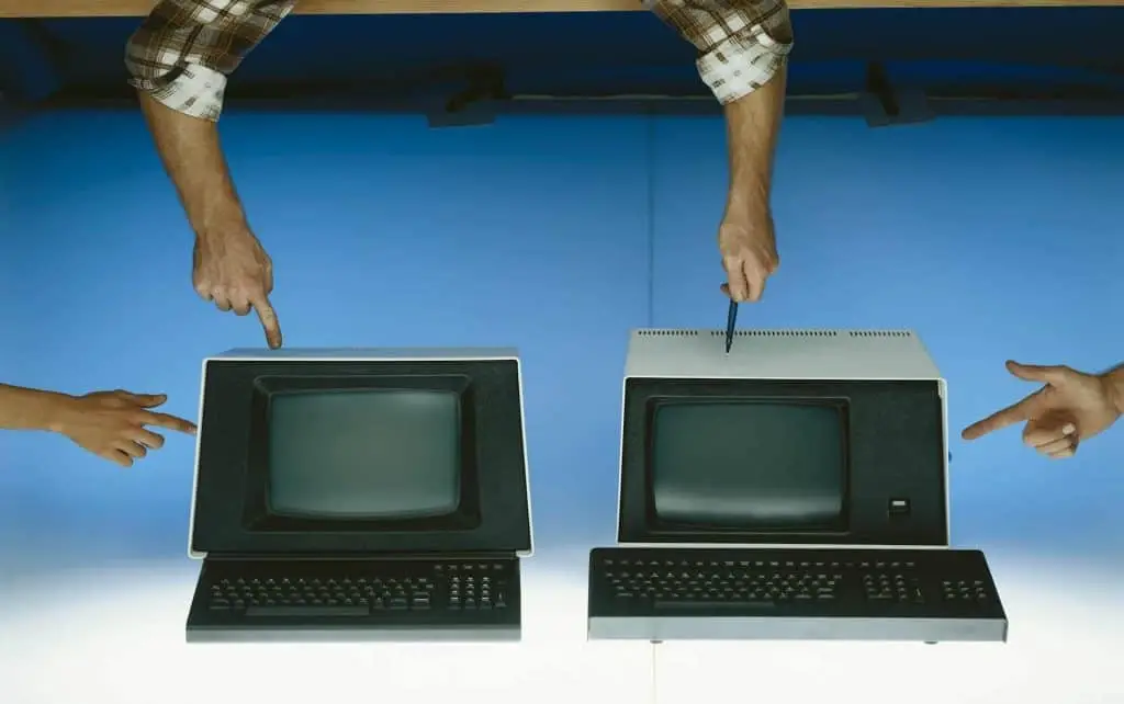 Two vintage computers are positioned side by side, with hands pointing at each one, against a blue background. Each monitor has a keyboard in front. These relics remind us of the growing pile of electronic waste in Atlanta, GA. Consider using a local drop-off box for proper disposal.