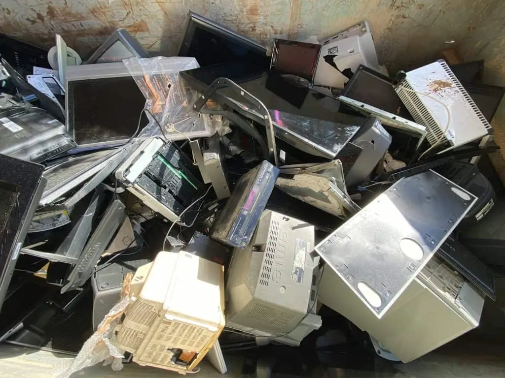 A pile of discarded electronics, including old computer monitors, towers, and partially dismantled devices, sits haphazardly in a drop-off box in Atlanta, GA. Sunlight highlights the various components and wires as part of the city's ongoing electronic waste management efforts.