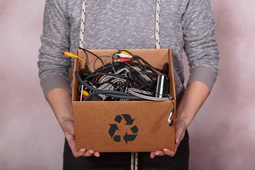 A person in a grey sweater holds a cardboard box filled with tangled cables and electronics, featuring a recycling symbol on the front. The background is neutral, highlighting the importance of electronic recycling in Atlanta, GA.