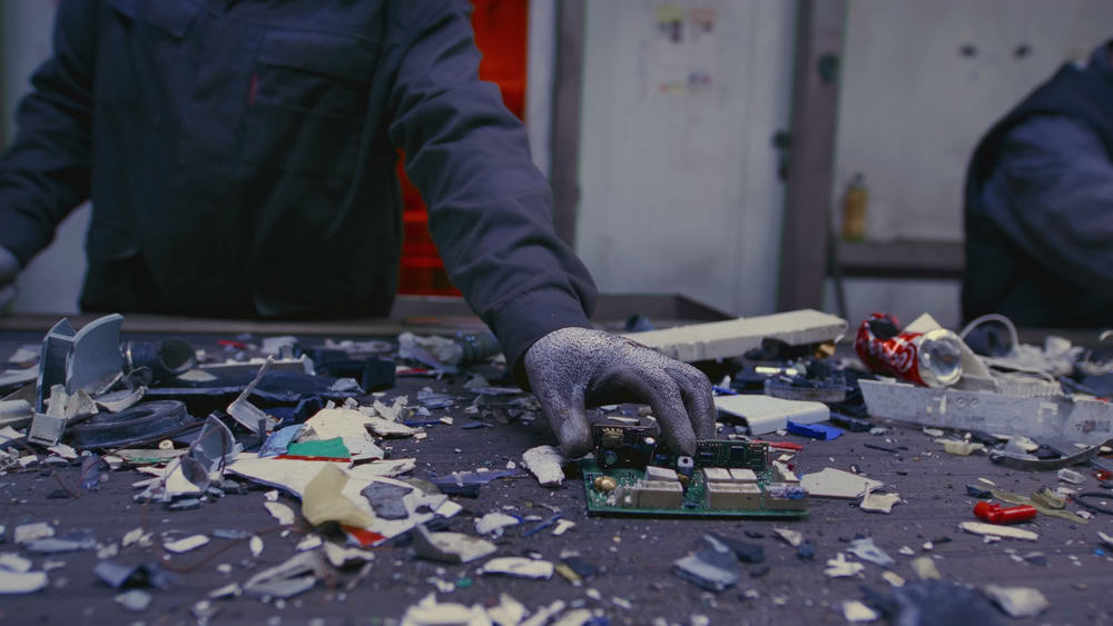 A person wearing a dark jacket and protective glove sorts through various electronic waste and components on a table. The scene suggests an e-waste disposal or recycling facility, where fragmented materials from recycled laptops and computers are scattered across the surface.