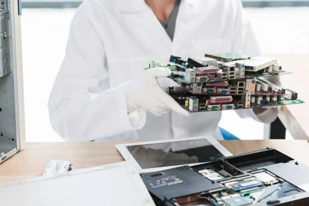 A person in a white lab coat and gloves holds several stackable computer circuit boards, poised for electronic recycling in Atlanta, GA. Nearby, electronic devices and disassembled parts are scattered on a wooden table in the bright workspace.