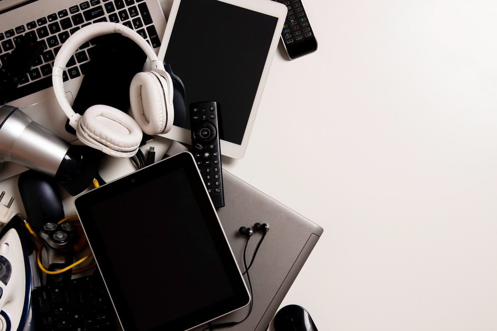 A cluttered desk with a laptop, tablets, headphones, a remote control, earbuds, a camera lens, an iron, and various cables scattered randomly on a white surface begs for electronics recycling. The right side of the image is empty.