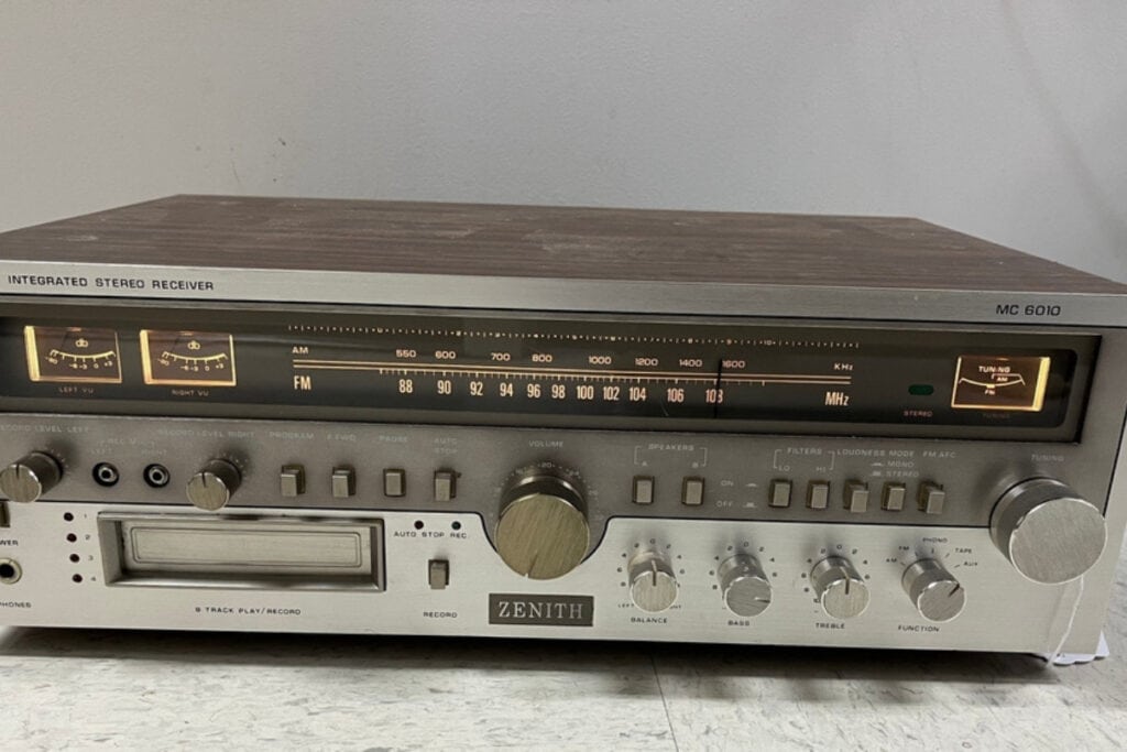 A vintage Zenith integrated stereo receiver, model MC 6010, with a wooden top shines on a light-colored surface. It features analog tuning dials for AM and FM radio, control knobs, buttons, and an 8-track player slot&mdash;perfect for those passionate about recycling electronics like old stereos.