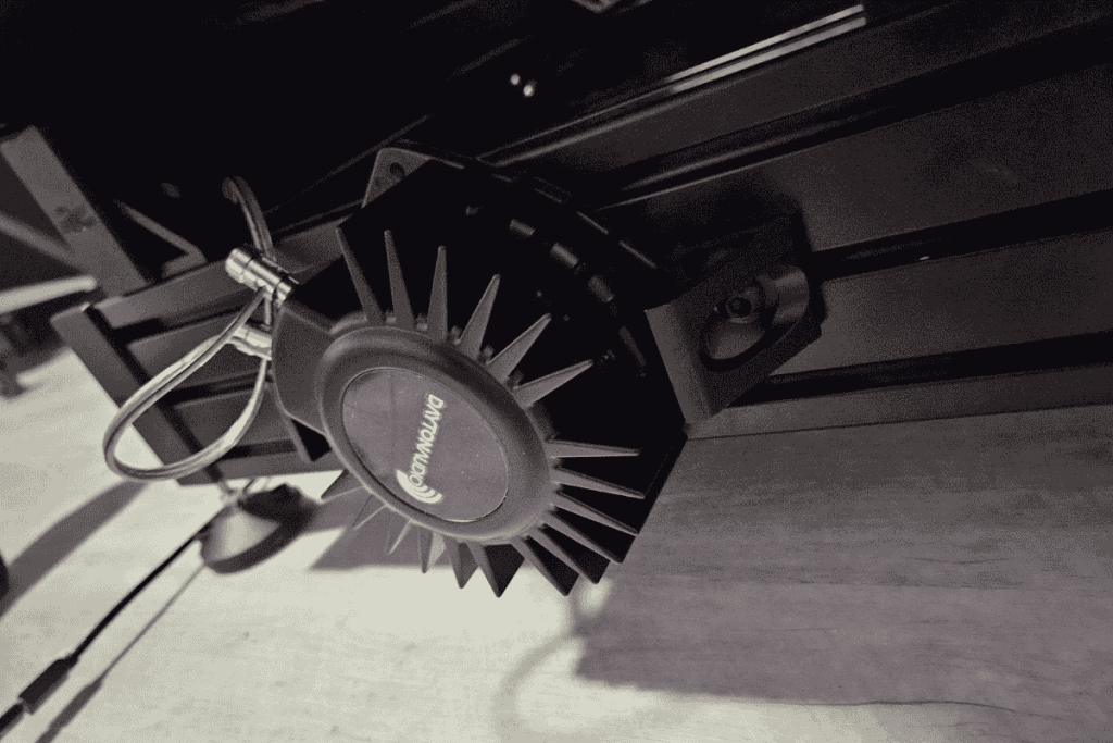 Close-up of a black machine component with metal fins, reminiscent of parts for recycle computers, mounted on a dark frame. Two wires are attached to the device. The brand "Anycubic" is visible on the front. The image is in black and white, highlighting the need for electronics waste disposal awareness.