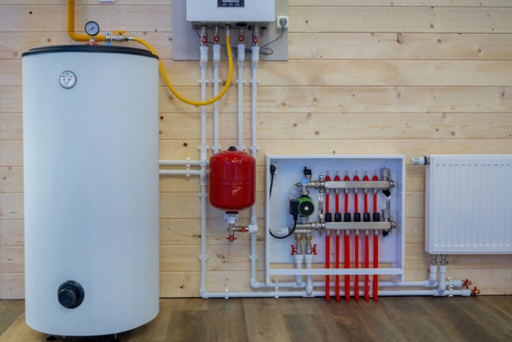 In a wooden-walled room, a modern heating system blends seamlessly with advanced technology. It features a large white tank, red expansion tank, control panel with gauges and knobs, various pipes, and a radiator on the right&mdash;demonstrating innovation akin to electronics recycling.