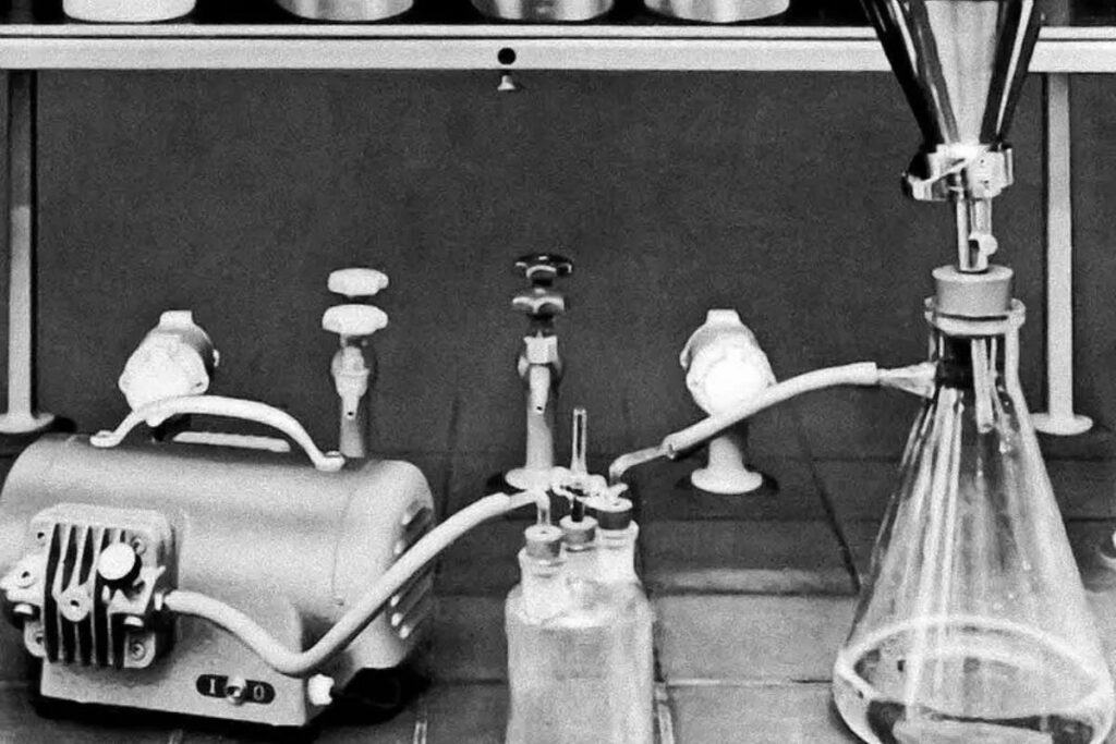 Black and white image of a laboratory setup with flasks, tubing, and a vacuum pump on a tiled surface. A large funnel is positioned above a flask on the right, hinting at sophisticated processes akin to electronics recycling. Valves and additional bottles adorn the background.