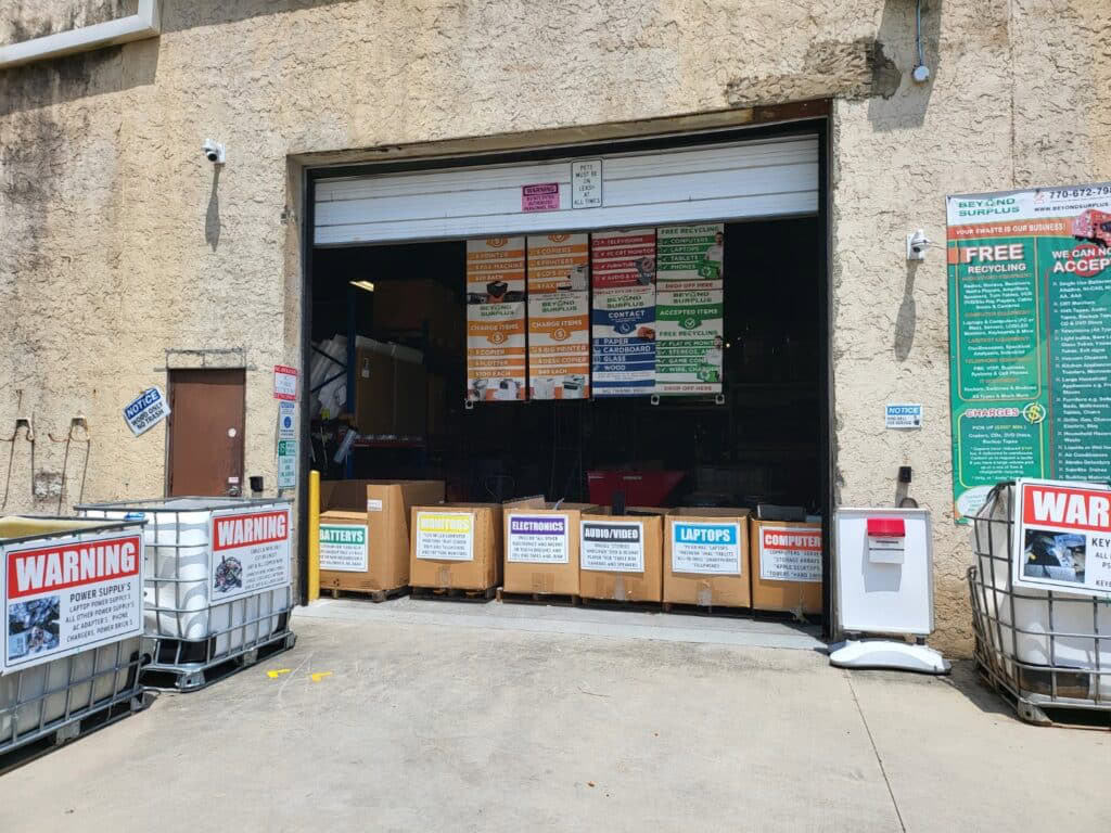 The warehouse entrance, with its partially open garage door, displays signs for electronics recycling. Warnings and drop-off categories such as batteries, recycle laptops, and computers are clearly marked. Large bins and information posters are prominently visible outside.