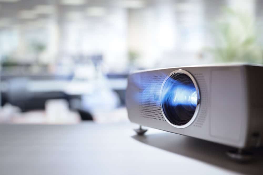 Close-up of a white projector on a table, emitting a beam of blue light. The softly focused office environment highlights the importance of electronics waste disposal, reminding us to recycle laptops and reduce e-waste in our workplaces.