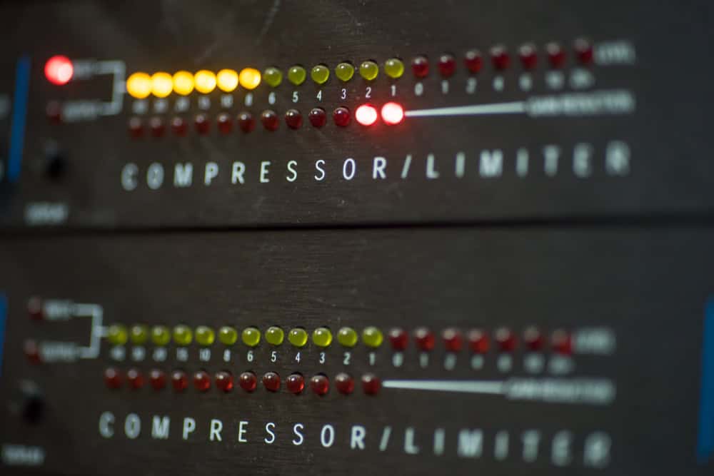 Close-up of a compressor/limiter audio device, reminiscent of finely tuned electronics ready for recycling, with rows of glowing LED lights in red, green, and yellow indicating audio levels. The device features dual displays that showcase varying degrees of audio compression or limiting.
