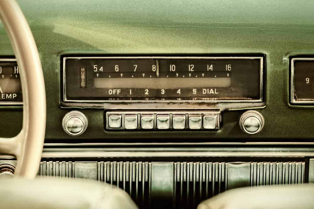 Close-up of a vintage car dashboard featuring an old-fashioned radio with push-button controls and numbered dials, reminiscent of eras before electronics recycling became crucial. The upholstery is a muted green color, and part of a steering wheel is visible on the left.