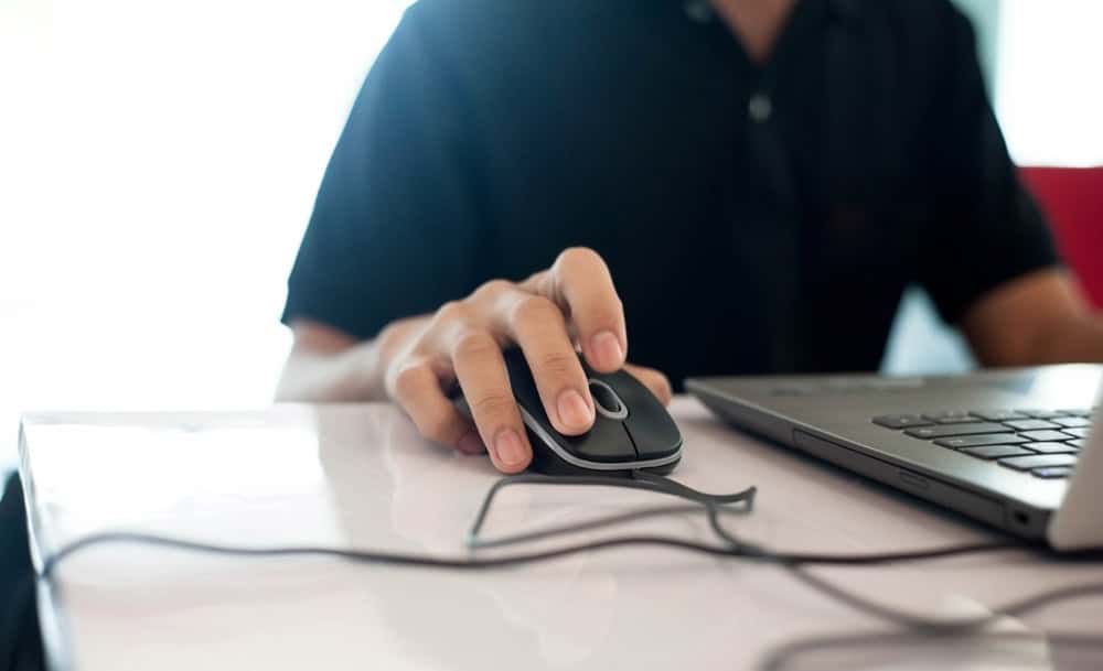 A person in a dark shirt uses a computer mouse next to a laptop on a white desk, mindful of electronics recycling. The focus is on the hand and mouse, with the laptop partially visible. The background is softly blurred, hinting at the importance of reducing electronics waste disposal.