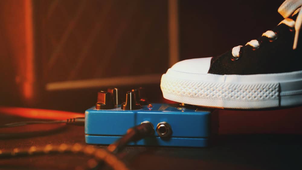 A close-up of a foot in a black and white sneaker pressing down on a blue guitar effects pedal, creatively repurposed from electronics waste. The pedal rests on a dimly lit stage, with an amplifier blurred in the background, reminding us to recycle laptops responsibly.