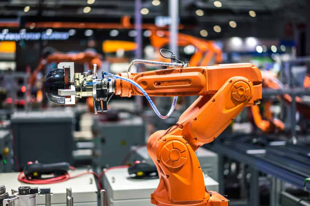 An orange robotic arm in a factory setting, equipped with wires and a gripper, efficiently handles electronics waste disposal. The blurred background of machinery and industrial equipment hints at a busy manufacturing environment focused on recycling computers and laptops.