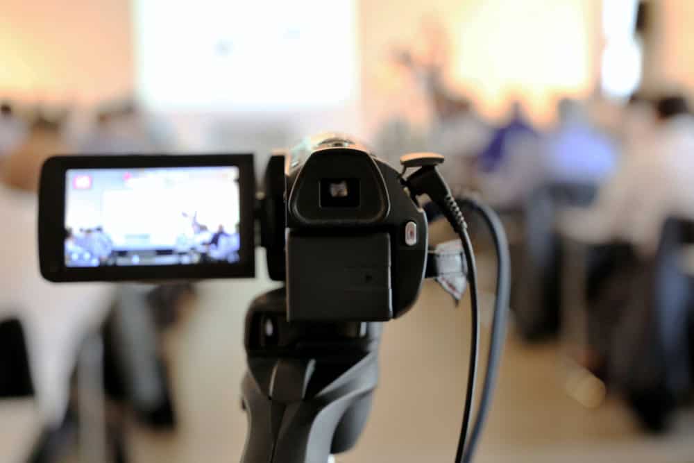 A video camera on a tripod is focused on a blurred audience in a conference room. The display screen shows an image of a projector presentation, highlighting the importance of electronics recycling at the front of the room. The scene is brightly lit and professional.