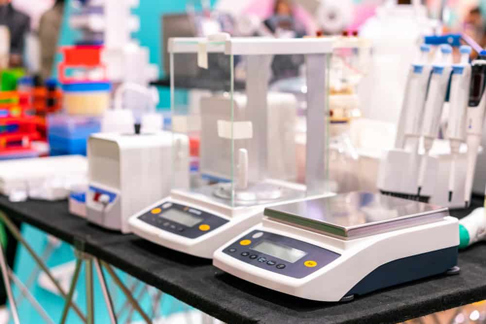 Laboratory equipment displayed on a table, including digital scales and pipettes, is neatly arranged. The setup subtly emphasizes the importance of electronics recycling in scientific settings, with a blurred background suggesting a scientific or exhibition setting.