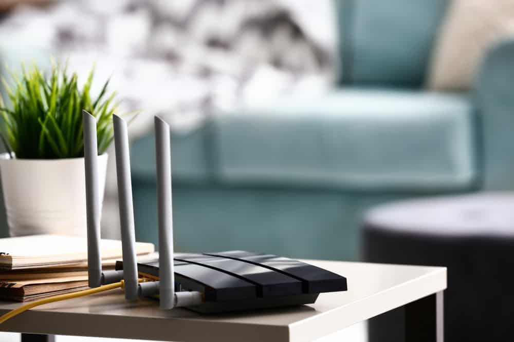 A black wireless router with three antennas sits on a white table, symbolizing the need for mindful electronics waste disposal. A yellow cable is plugged in. In the background, there's a potted plant, a stack of notebooks, a light blue sofa, and a blurred cushion.