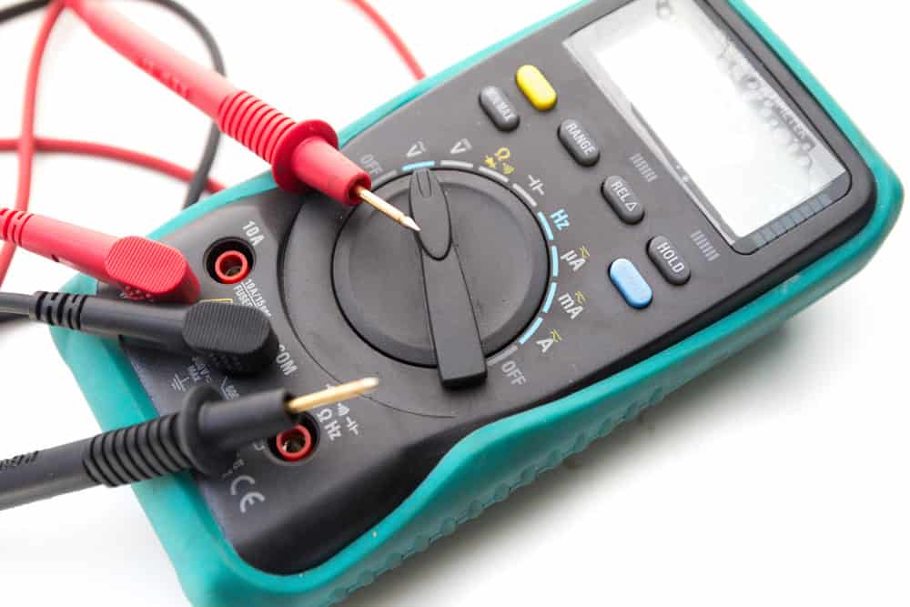 A digital multimeter with black and red probes is shown, set to measure resistance, as indicated by the dial pointing to the "&Omega;" symbol. Housed in a green protective casing, it's a reminder of the importance of proper electronics waste disposal for sustainable tech use.