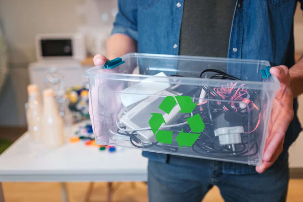 A person holds a transparent plastic container filled with various electronic cables and devices, symbolizing electronics recycling. A green recycling symbol is displayed on the box. In the background, a table with plastic bottles and caps highlights the commitment to responsible waste disposal.